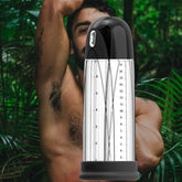 A person with a beard and arm raised stands partially obscured by a large transparent cylindrical object—VeDO's Pump Rechargeable Vacuum Penis in 'Just Black'. The background features lush green leaves, creating a natural jungle-like setting, suggesting enhanced stamina and vitality.